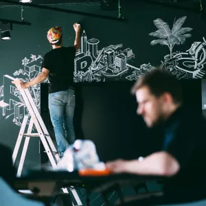 Person stands on ladder and draws detailed illsutration with white acrylic pens on a dark green wall, a second person plans the next contents of the wall graphic on a laptop in the foreground