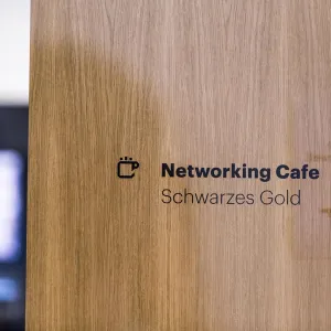 Signage consisting of icon and sans serif font in black on a glass pane, the sign shows a coffee cup and the text: Networking Cafe Schwarzes Gold