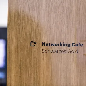Signage consisting of icon and sans serif font in black on a glass pane, the sign shows a coffee cup and the text: Networking Cafe Schwarzes Gold