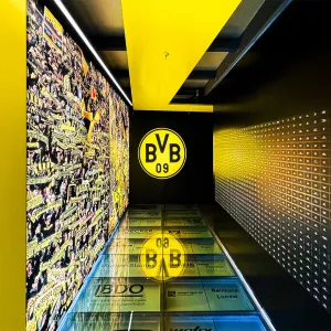 Gang im Borusseum zeigt auf der linken Wand ein flächeneckendes Foto einer BVB Fankurve, auf der rechten Seite befinden sich Kacheln mit Plaketten aller aktuellen und ehemaligen Spieler, im Boden eingelassen unter Glaskacheln sichtbar sind die Vereinssponsoren