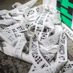 Weiße Papierstreifen mit schwarzem Text liegen auf grauem Untergrund verstreut, neben einem weißen Behälter mit grünen und magentafarbenen Elementen. Die Streifen scheinen Teil einer Ausstellungsinstallation zu sein und tragen Teile eines langen Manifests.