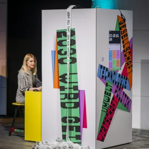 Eine Frau sitzt neben einer großen weißen begehbaren Installationen mit bunten Texten und Grafiken in Grün, Orange, Magenta und Blau. Die Struktur ähnelt einem geöffneten Buch oder einer dreidimensionalen Ausstellungswand in einem modernen Ausstellungsraum mit dunklem Hintergrund und blauer Beleuchtung.