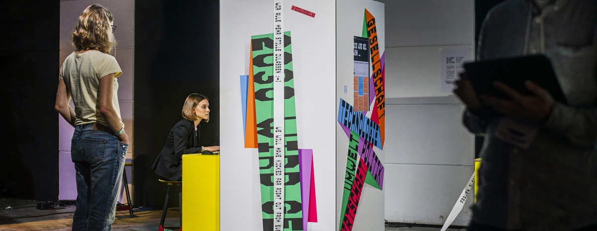 Ein weiße Ausstellungsinstallation mit bunten, farbenfrohen Grafiken und Texten steht in einem dunklen Ausstellungsraum. Mehrere Besucher erkunden die Installation von verschiedenen Seiten, während einer von ihnen ein Mobilgerät hält. Gelbe Elemente und Kabel ergänzen die Ausstellungsgestaltung.