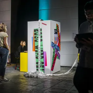 Eine weiße Ausstellungsinstallation mit bunten Wort-Fragmenten steht in einem modernen Ausstellungsraum. Besucher betrachten die bedruckte Struktur, an der bunte Papierstreifen herunterhängen. Eine gelbe Säule und moderne Wandbeleuchtung rahmen die kunstvolle Darstellung ein.