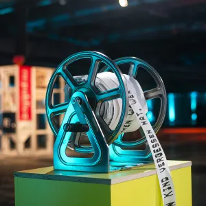 Eine bedruckte Papierrolle stehen auf einem gelben Sockel in einer modernen Ausstellung. Im unscharfen Hintergrund sind bunte LED-Lichter und Ausstellungselemente zu sehen.