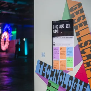 Ausstellungsansicht mit bunten Wandgrafiken in Grün, Orange, Rosa und Blau sowie typografischen Elementen zum Thema Technologie. Links im Hintergrund ein Ausstellungsbereich mit leuchtenden Neon-Objekten und farbigen Lichtern. Rechts an der Wand ein großes Manifest-Installation mit mehrfarbigen Textblöcken und schräg angeordneten Buchstaben.