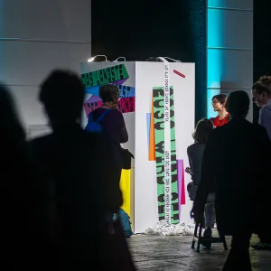 Besucher interagieren mit einer großen weißen interaktiven Installation in einem dunklen Ausstellungsraum mit blauer Beleuchtung. Die bunte Wand zeigt Texte und Grafiken und wird von mehreren Menschen erkundet, die vor ihr stehen und sich damit beschäftigen.