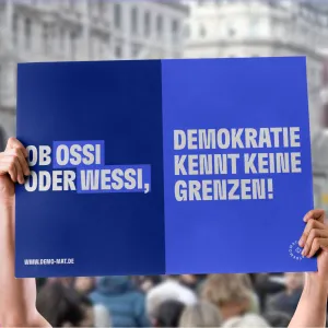 Demo-Mat messages at demos, the messages can be easily shared and printed out. A poster held up with a blue background reads: Whether Ossi or Wessi, democracy knows no borders!