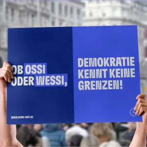 Demo-Mat Botschaften auf Demos, die Botschaften lassen sich einfach Teilen und Ausdrucken. Auf einem hochgehaltenen Poster mit blauem Hintergrund steht: Ob Ossi oder Wessi, Demokratie kennt keine Grenzen!