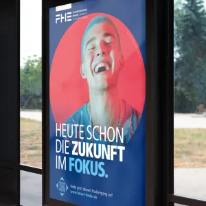 A portrait format poster hanging inside a bus stop. The motif consists of a young man with light blue hair in a red circle on a dark blue background. The campaign headline can be read again in the foreground.