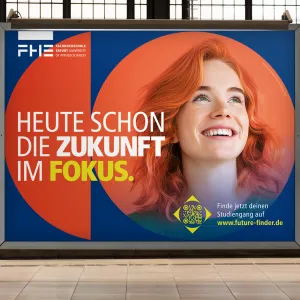 A billboard on a platform, with a train on the track in the background. The main motif can be seen on the poster. The red-haired woman with an orange background in a geometric shape on a dark blue background. Above it is the headline of the first campaign phase. Focusing on the future today.