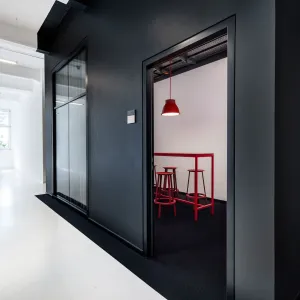 The black-painted wall with the entrances to the meeting rooms contrasts with the surrounding white walls, an open door to a meeting room shows friendly, bright meeting rooms, with a small glass front and red lampshades, a red high table and red bar stools