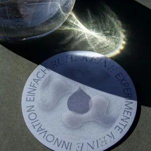 Round light blue glass coaster with a water droplet design in the center and the text “No experiments, no innovations, just RUH” running along the edge