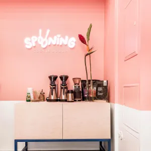Coffee station on a wooden cabinet below a glowing Spooning logo in front of a pink wall
