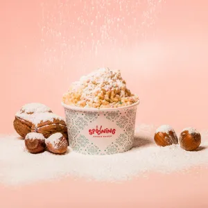 A paper cup well filled with cookie dough, printed with the Spooning Cookie Dough logo and pattern, nuts lying next to the cup and powdered sugar trickling down from above, covering the whole scene like snow